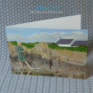 Art of Ireland greeting card of a woman in a green coat, standing on the Irish coast with her foot pointed toward a stone.
