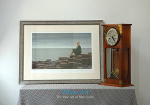 Framed Art Print sitting on a table showing an oil painting of a lady seated on the Giant's Causeway on an overcast day.