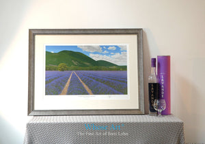 Framed art print of a lavender field painting set in the south of France. The Framed art print is resting on a table by a lamp