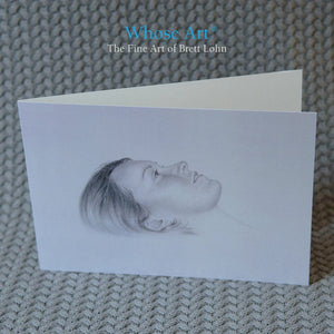 Pencil greeting card of a drawing of the head of a young woman who is daydreaming. The greeting card stands on a table